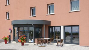 Fachada de hotel con entrada curva, zona de estar exterior con mesas y sillas, y amplias ventanas.