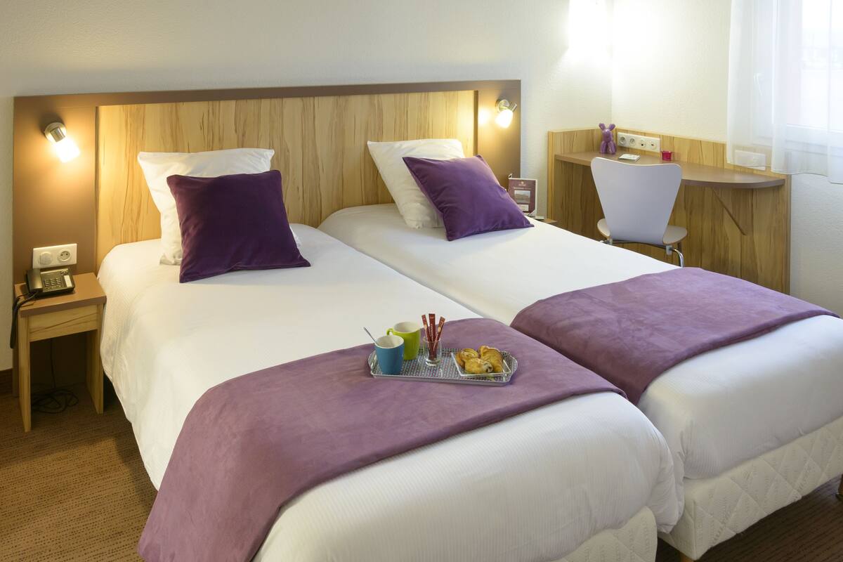 Ein Hotelzimmer mit zwei Betten, einem Schreibtisch und einem Stuhl. Auf dem Bett steht ein Tablett mit Speisen und Getränken.