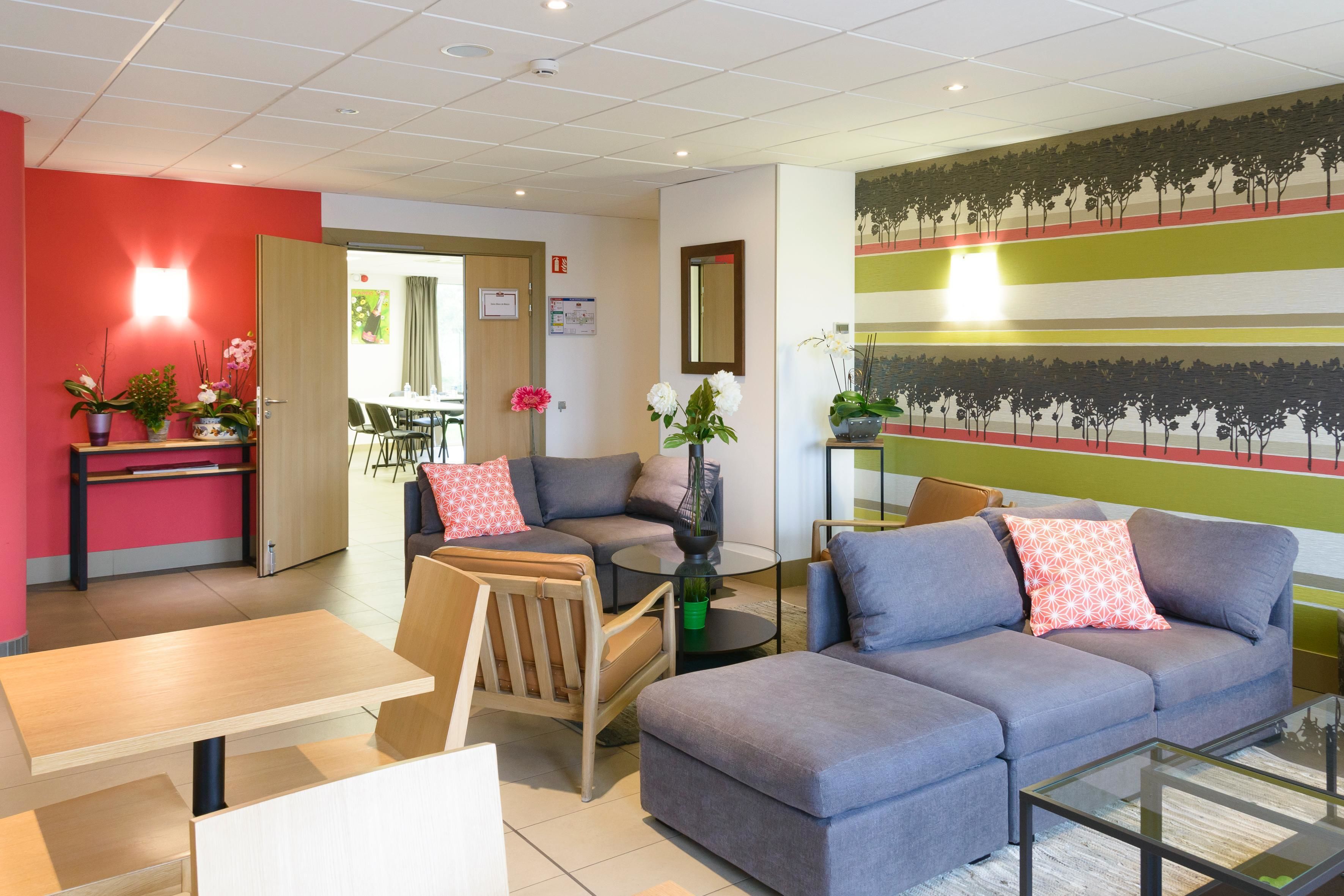 En moderne hotellobby med en grå sofa, en træstol, et glastæppe og et lille bord med stol. Der er krukkeplanter og en væg med et træ-motiv.