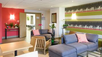 En moderne hotellobby med en grå sofa, en træstol, et glastæppe og et lille bord med stol. Der er krukkeplanter og en væg med et træ-motiv.
