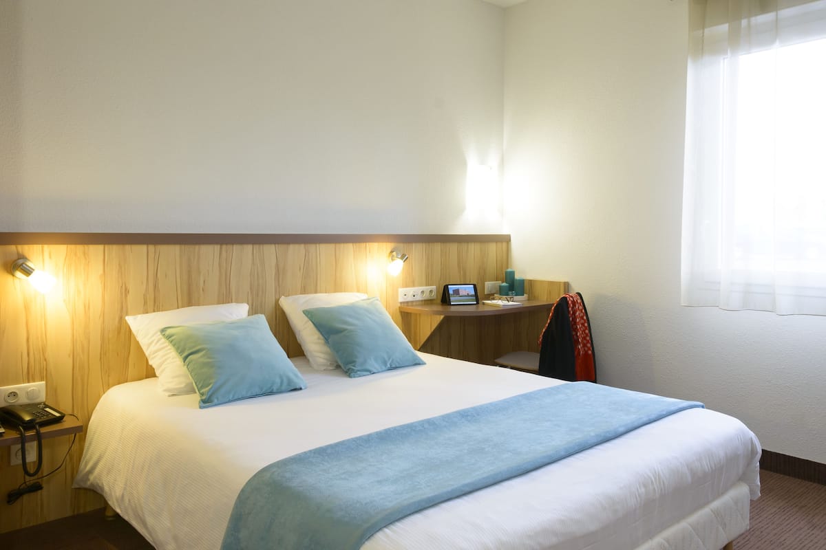 Ein Hotelzimmer mit einem großen Bett, einem Nachttisch mit Lampe, einem kleinen Schreibtisch mit Computer und einem Fenster mit Vorhängen.
