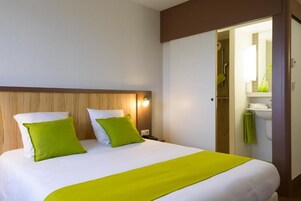 A hotel room with a bed, a nightstand, a wall-mounted lamp, and a bathroom visible through an open door.