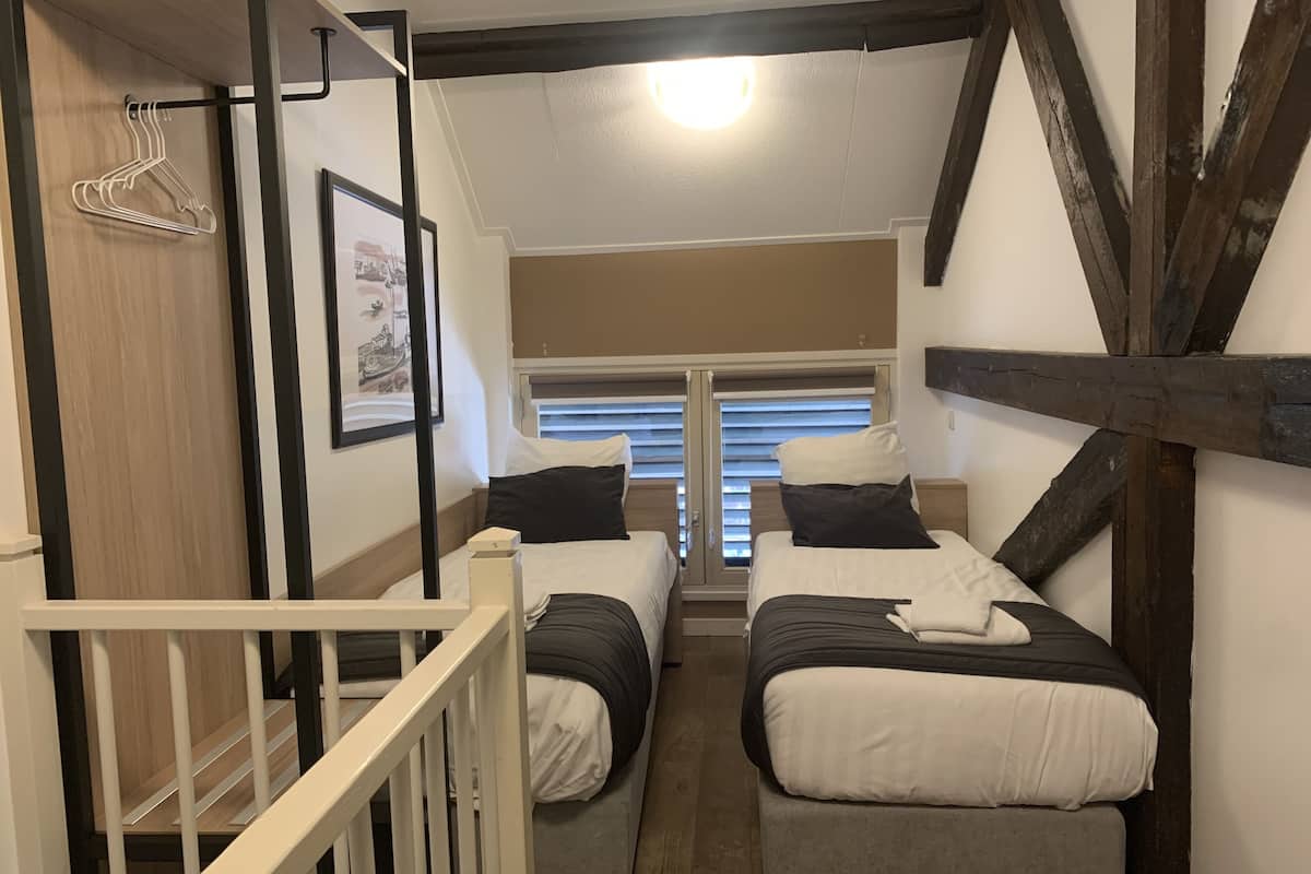 A bedroom with two single beds, a wooden ceiling, a window with blinds, and a white railing.