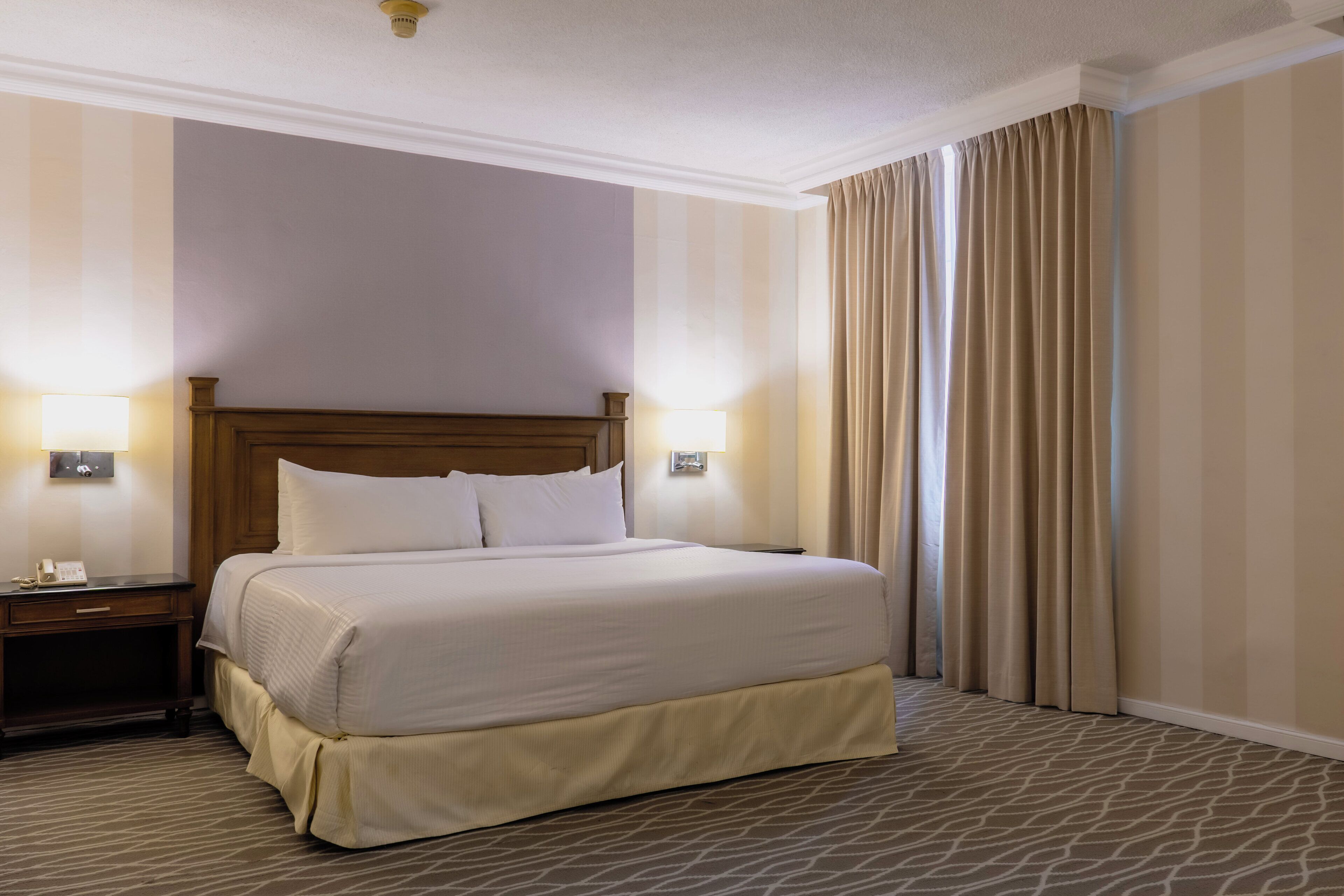 A hotel room with a large bed, two bedside tables, a nightstand, and a window with curtains.