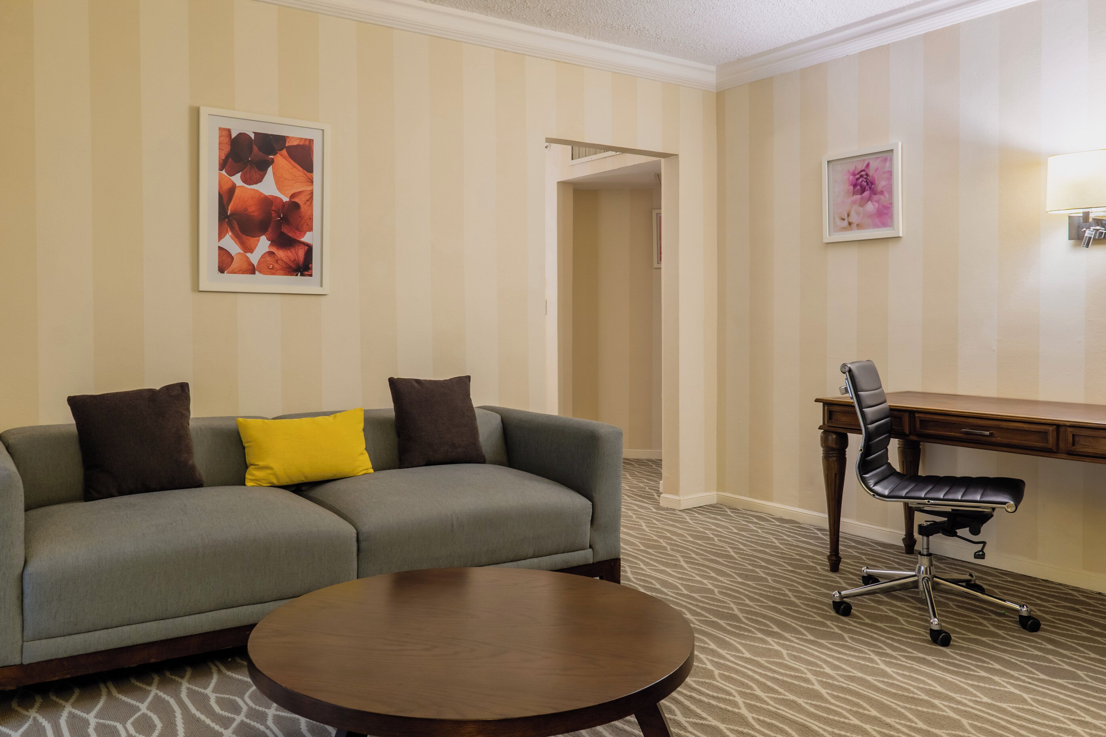 Una habitación de hotel con un sofá gris, una mesa redonda de madera, un escritorio con una silla de oficina negra y papel tapiz a rayas.