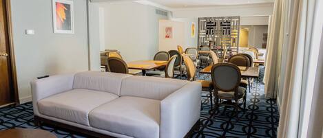 A hotel room with a sofa, dining table, chairs, and a patterned carpet.