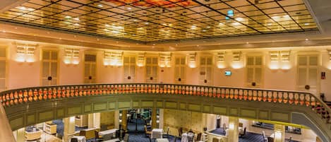 Vestíbulo de hotel de varios niveles con piso de diseño ajedrezado, techo de vidrio y un balcón curvo con asientos.