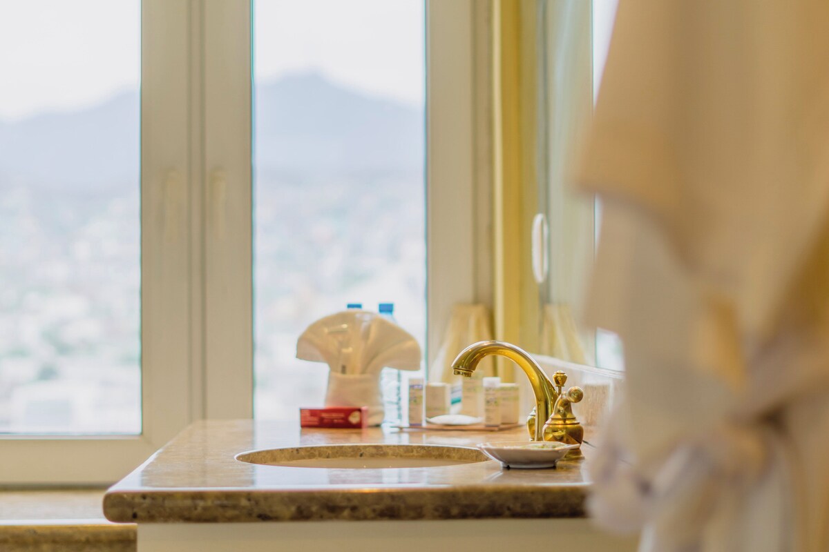 Un baño con lavamanos, un grifo dorado y una ventana que ofrece vistas a un paisaje urbano.