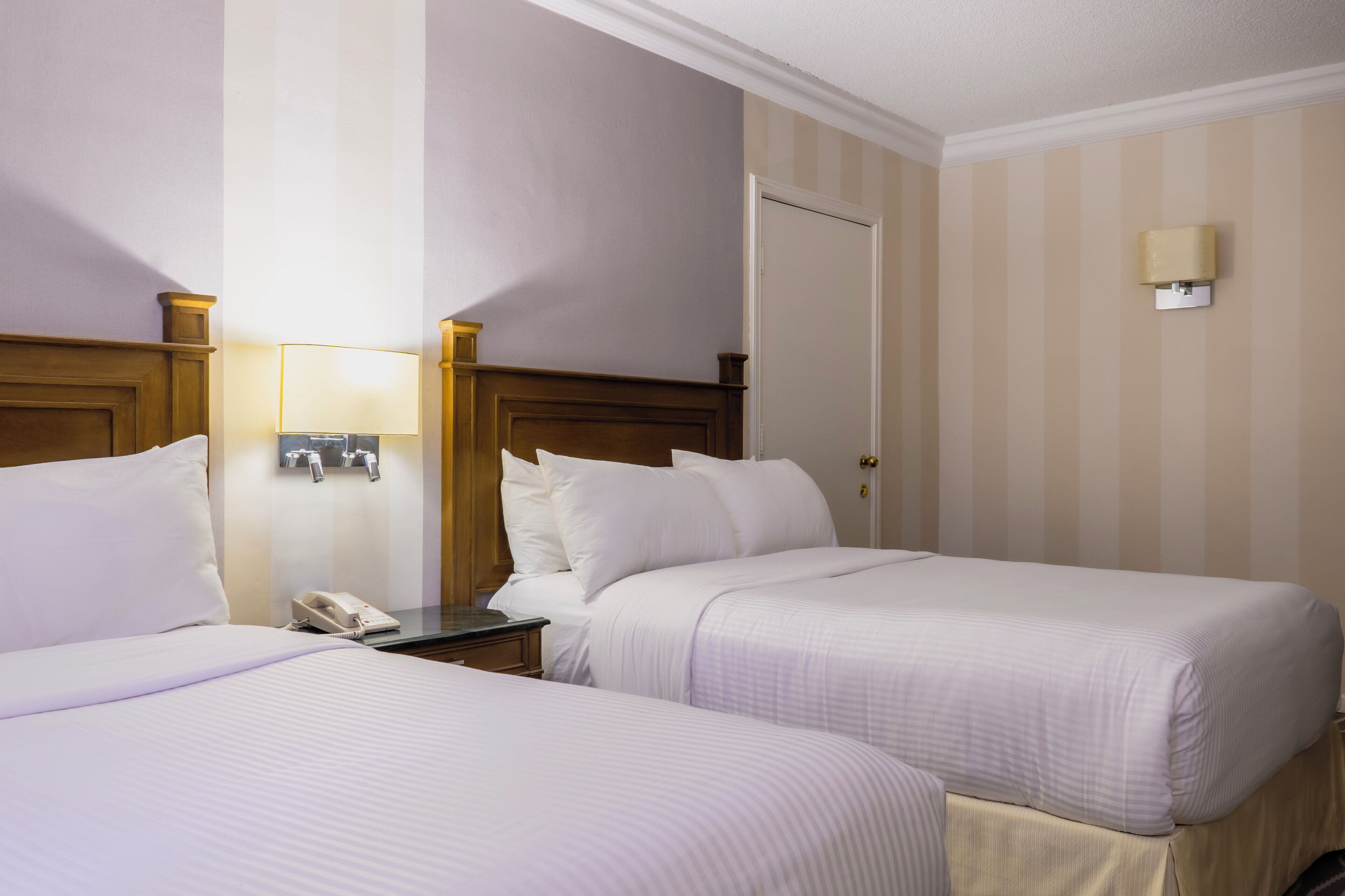 A hotel room with two beds, a nightstand with a phone, and a wall-mounted light fixture.