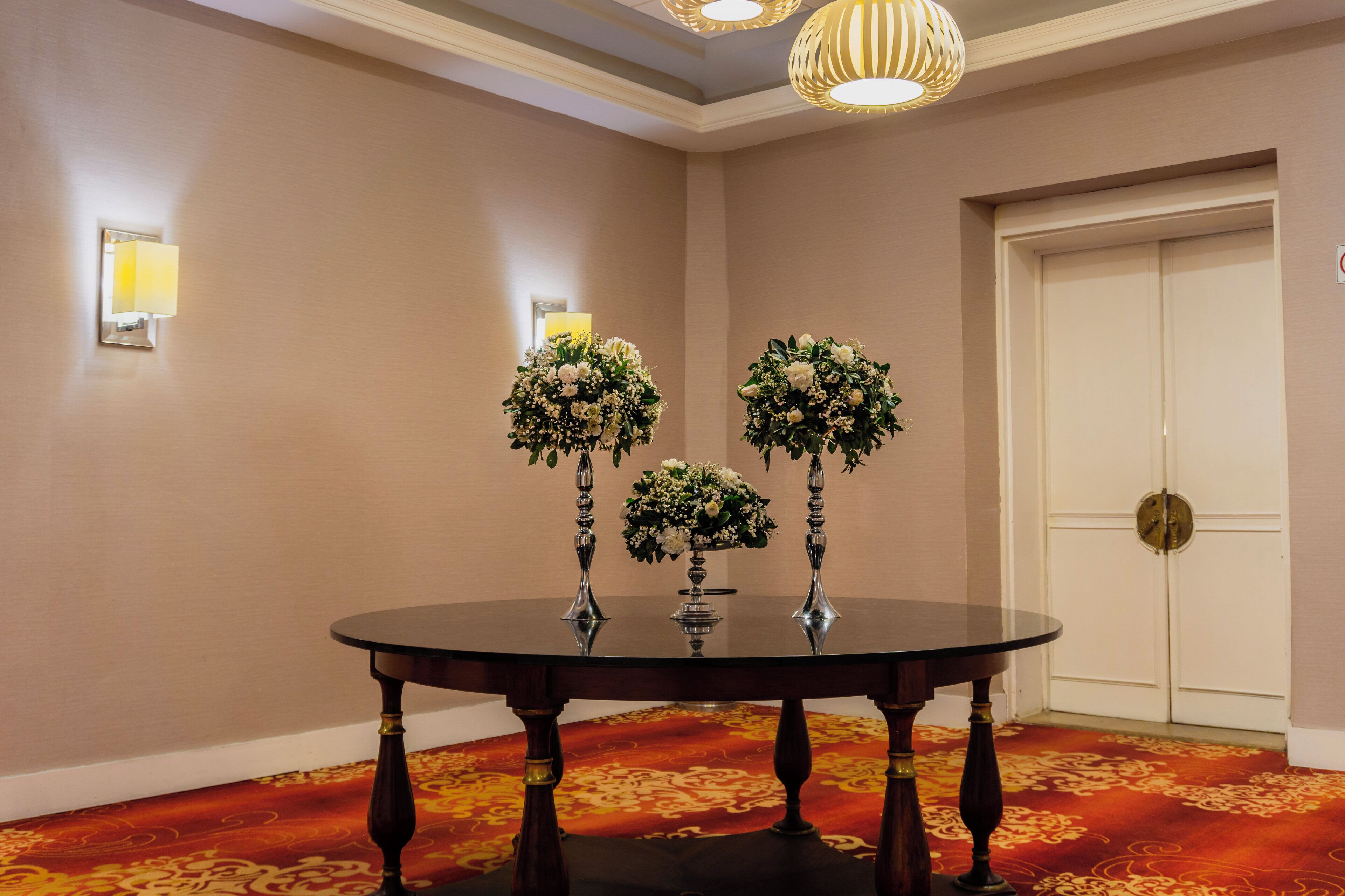 Une table ronde ornée de trois vases de fleurs, un tapis à motifs et une porte visible au fond.