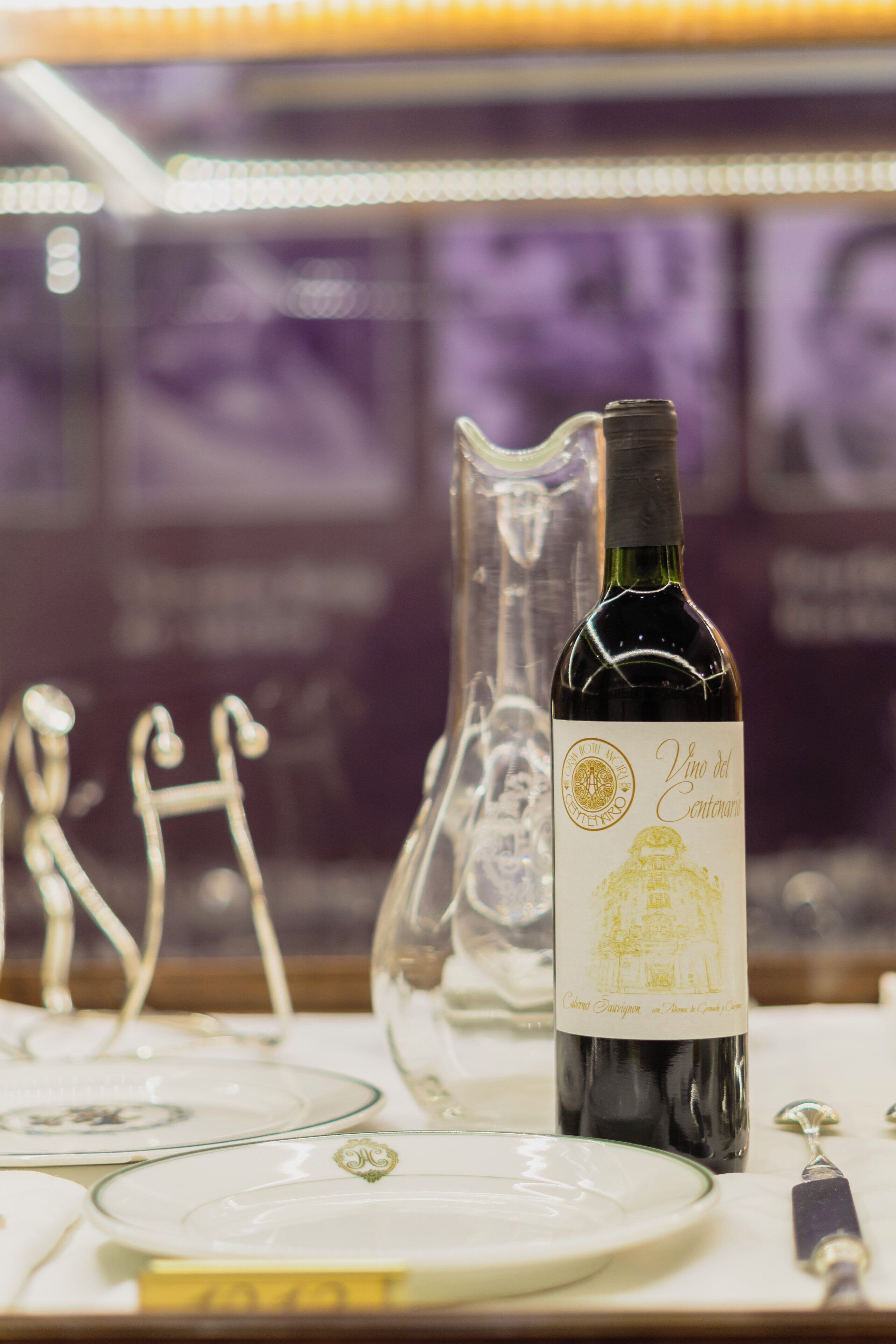 Una botella de vino con etiqueta, sobre una mesa con una jarra de vidrio y una figura metálica decorativa.