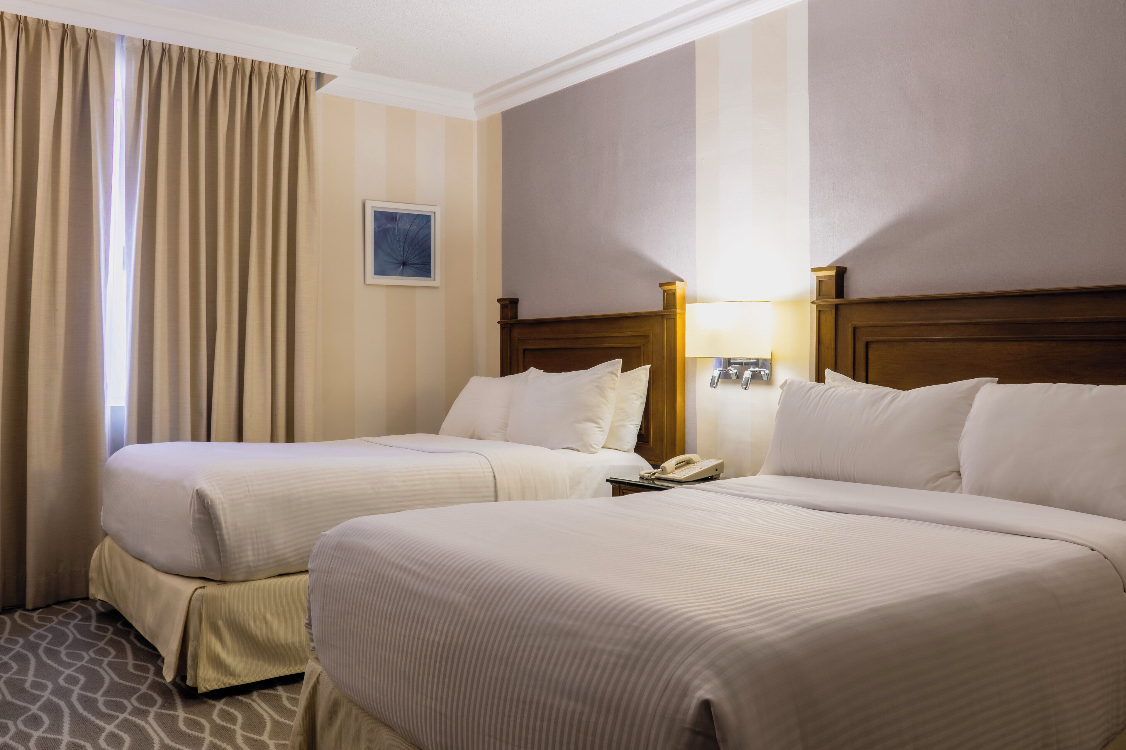 A hotel room with two beds, a bedside table with a lamp, a telephone, and a window with curtains.
