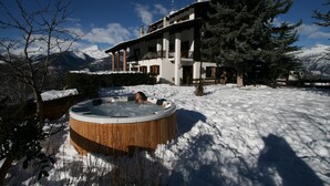 A wooden hot tub in a snowy landscape with a large house and mountains in the background. - Hotel Tivet (Gressan)