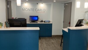 Un espace d’accueil d’hôtel au design moderne, comprenant un comptoir d’accueil, un écran d’ordinateur et un panneau mural indiquant « Promenade de MÔNTE-CARLO ».
