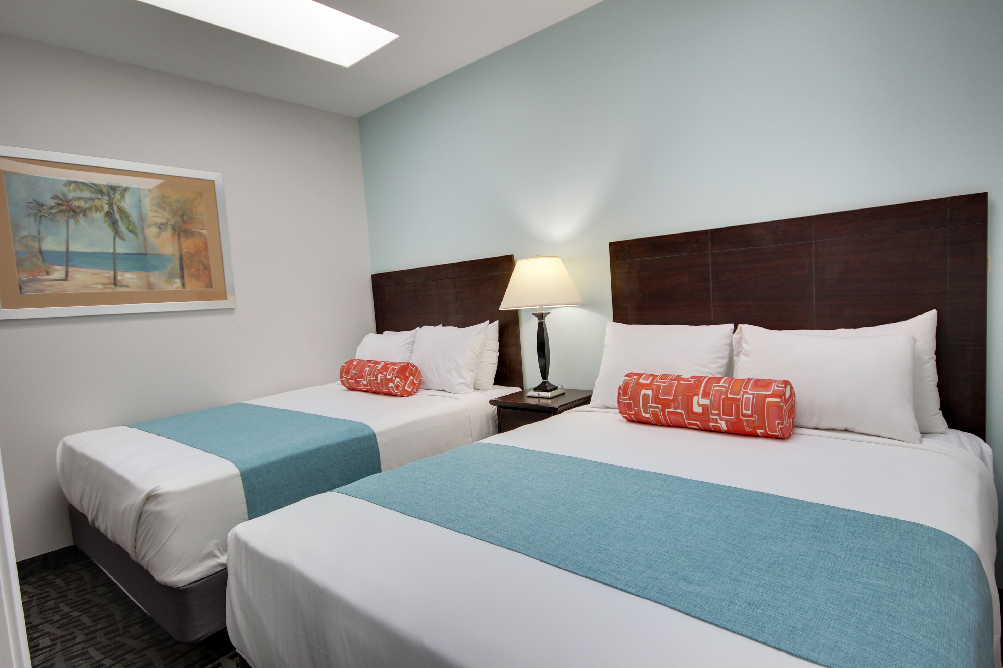 Ein Hotelzimmer mit zwei Einzelbetten, beide mit blauer Bettdecke und weißen Kissen. Dazwischen steht ein Nachttisch mit einer Lampe, und an der Wand hängt ein Palmenbild.