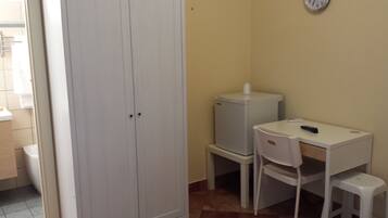 A small room with a white wardrobe, a small white desk with a chair, a white stool, and a white refrigerator.