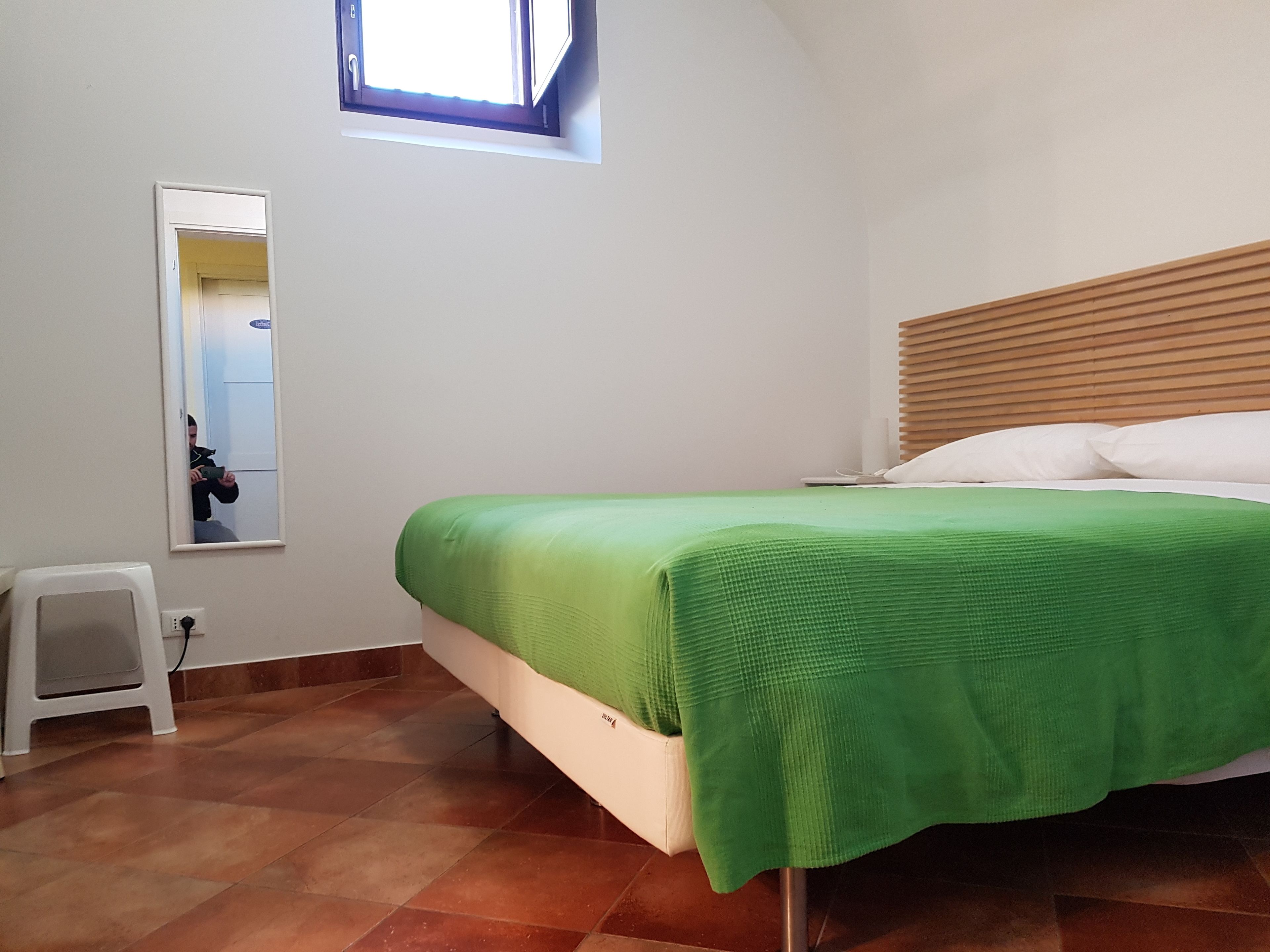 Ein Schlafzimmer mit einem Bett, einem kleinen Hocker, einem Spiegel und einem Fenster.