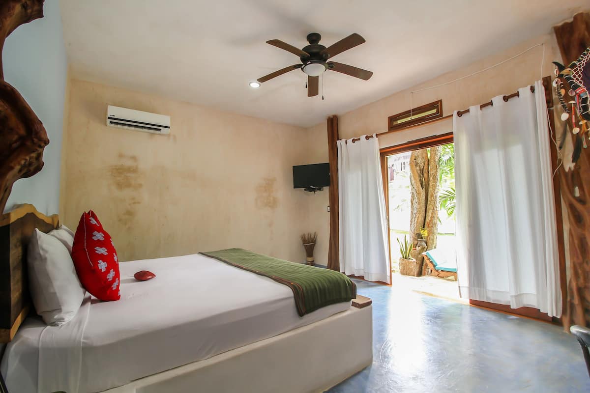 A bedroom with a bed, an air conditioner, a ceiling fan, a TV, and a view of the outdoors.