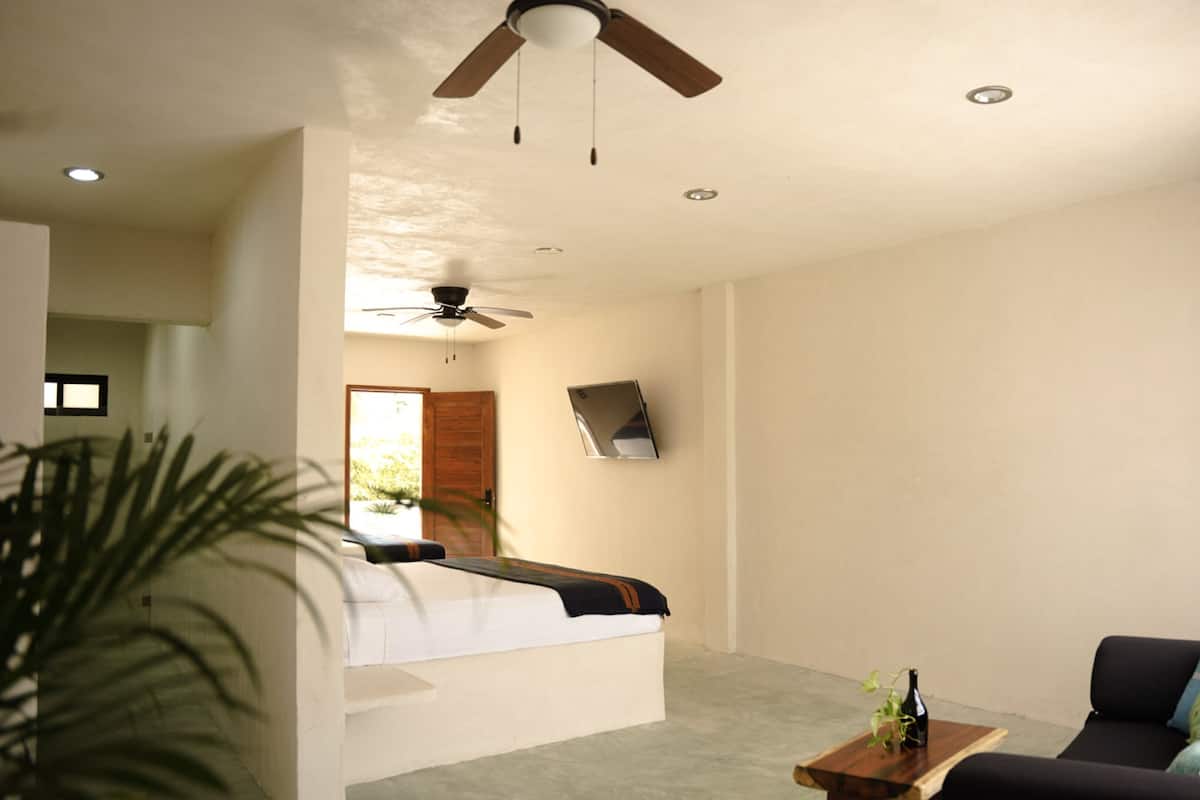 A modern bedroom with a bed, a ceiling fan, a small table with a bottle, and a sofa with pillows.