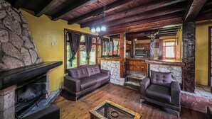 Ein rustikaler Innenraum mit Kamin aus Stein, Holzbalkendecke und Steinwand. Zwei Ledersofas und ein Couchtisch aus Holz sind ebenfalls vorhanden.