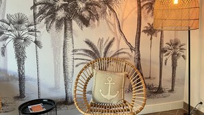 A room with a mural of palm trees, a wicker chair with an anchor pillow, a floor lamp, and a small round table with a book. - Hôtel de la Côte Fleurie (Deauville)
