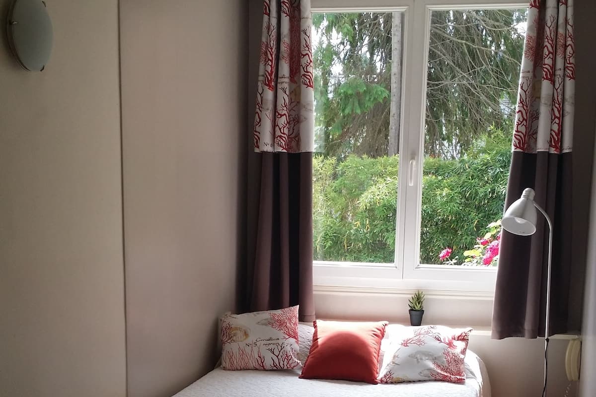 Ein Schlafzimmer mit einem Bett, einem Sessel, einem Nachttisch, einer Lampe und einem Fenster mit Vorhängen.