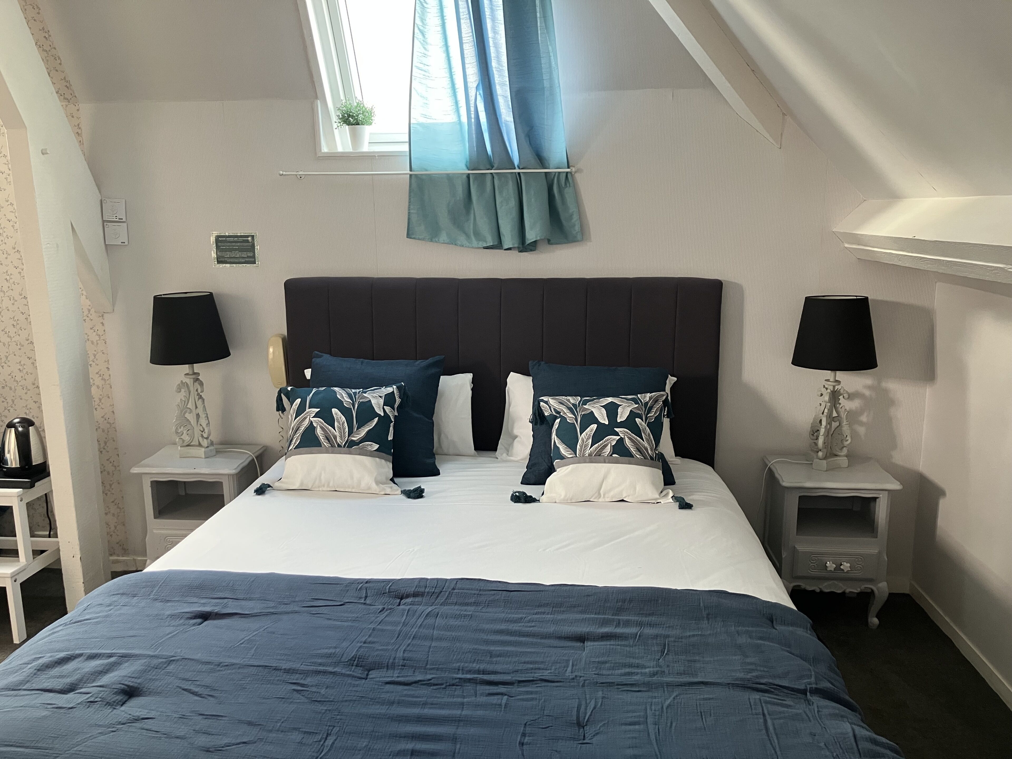 A neatly made bed with a dark blue bedspread and matching pillows, flanked by two bedside tables with lamps, in a room with a sloped ceiling and a window with blue curtains.