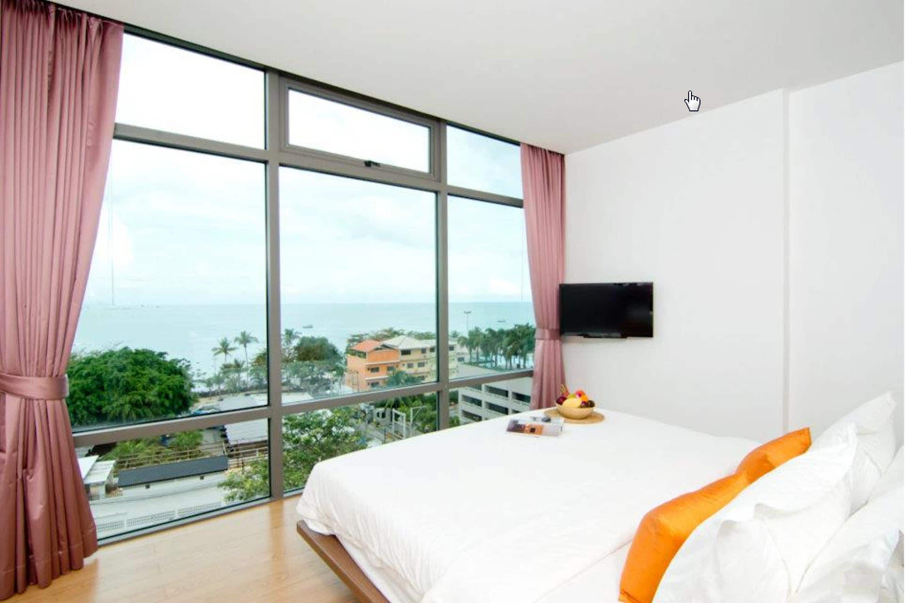 A hotel room with a large window, a bed with white bedding and orange pillows, a flat-screen TV on the wall, and a fruit bowl on the bed.