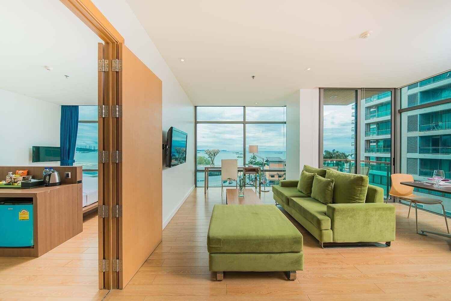 A modern hotel room with a green sofa, a wooden desk, a TV, and large windows offering a view of the city and the sea.