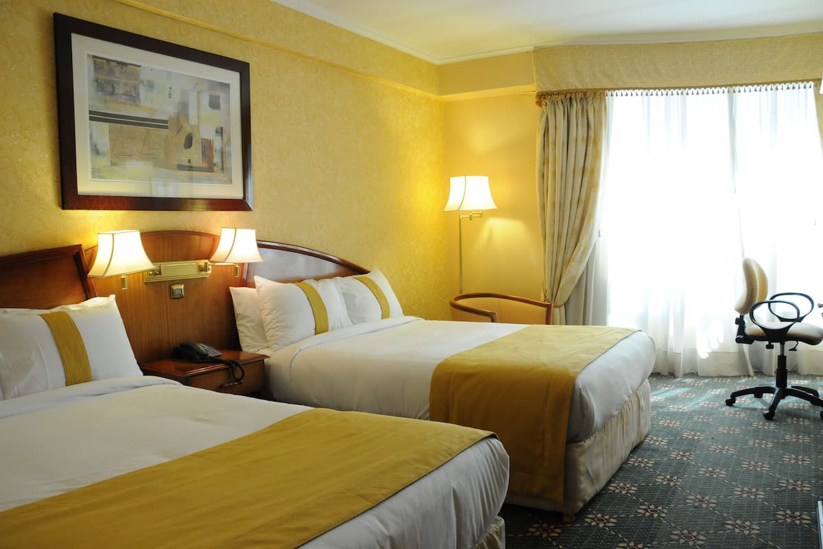 A hotel room with two beds, a desk, a chair, and a lamp.