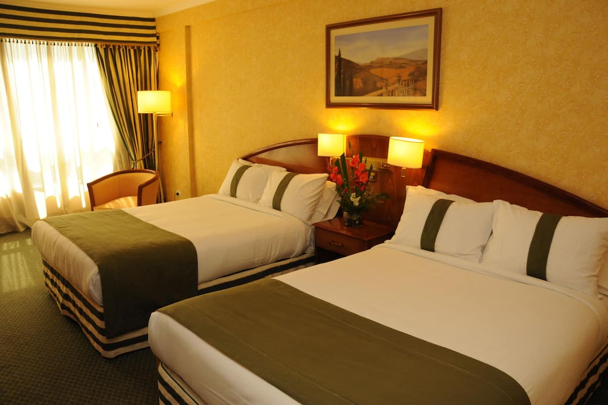 A hotel room with two beds, a nightstand with lamps, a chair, and a painting on the wall.
