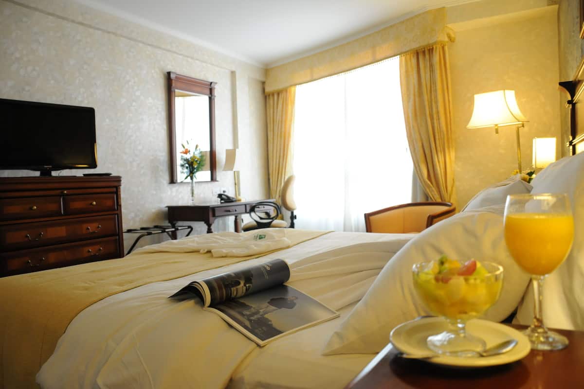 A hotel room with a bed, a television, a desk with a lamp, and a window with curtains.