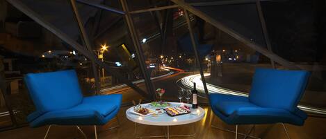 Un interior moderno con dos sillas azules alrededor de una mesa redonda, vistas a la ciudad a través de amplios ventanales y un paisaje urbano nocturno visible en el exterior.