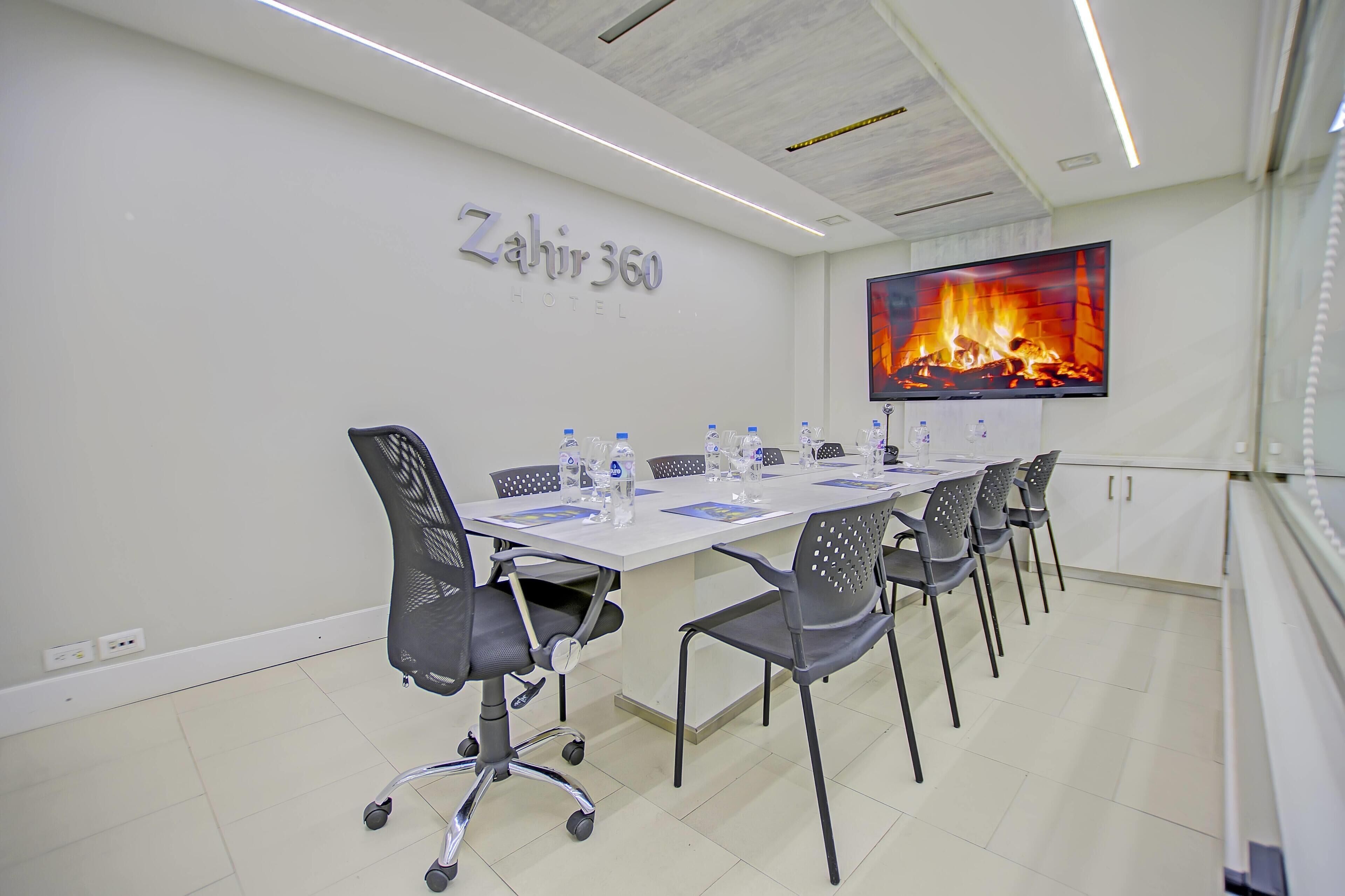 A conference room at Zahir 360 Hotel with a long table, chairs, a flat-screen TV, and a fireplace image on the wall.