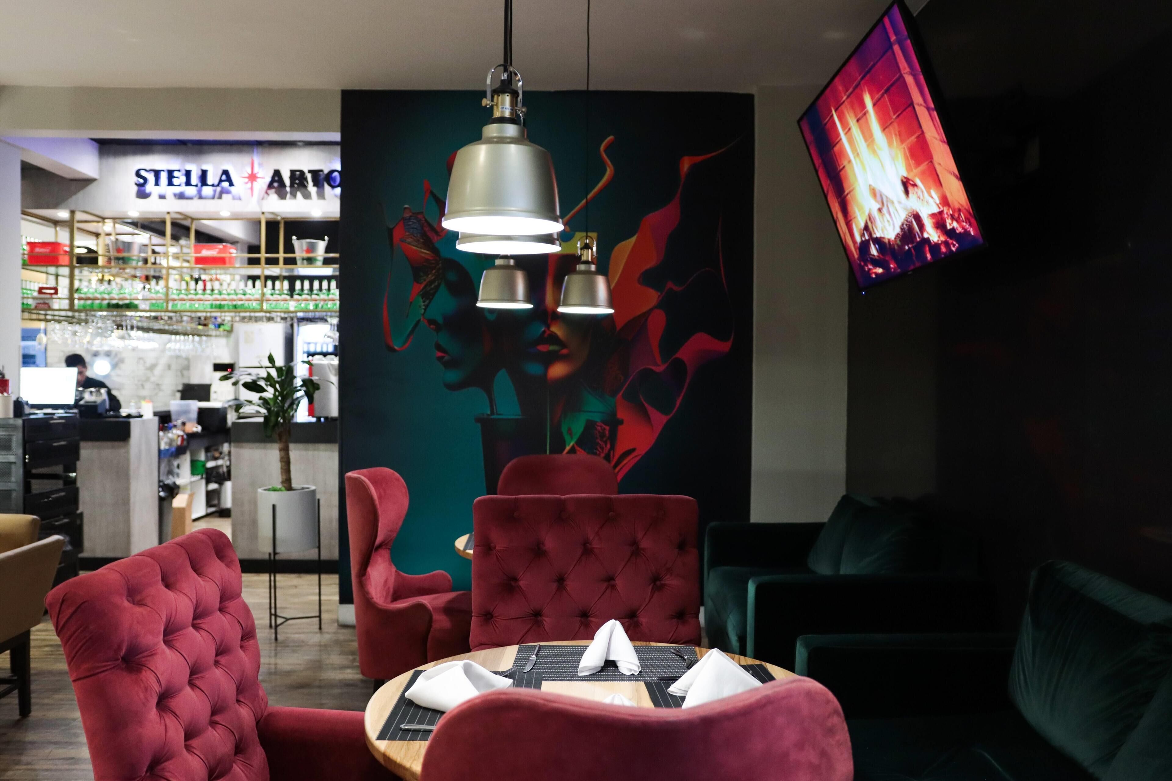 Un interior moderno con sillas tapizadas de rojo, una mesa redonda con servilletas, una zona de bar con un letrero 'STELLA ARTO' y una pantalla grande mostrando un fuego.