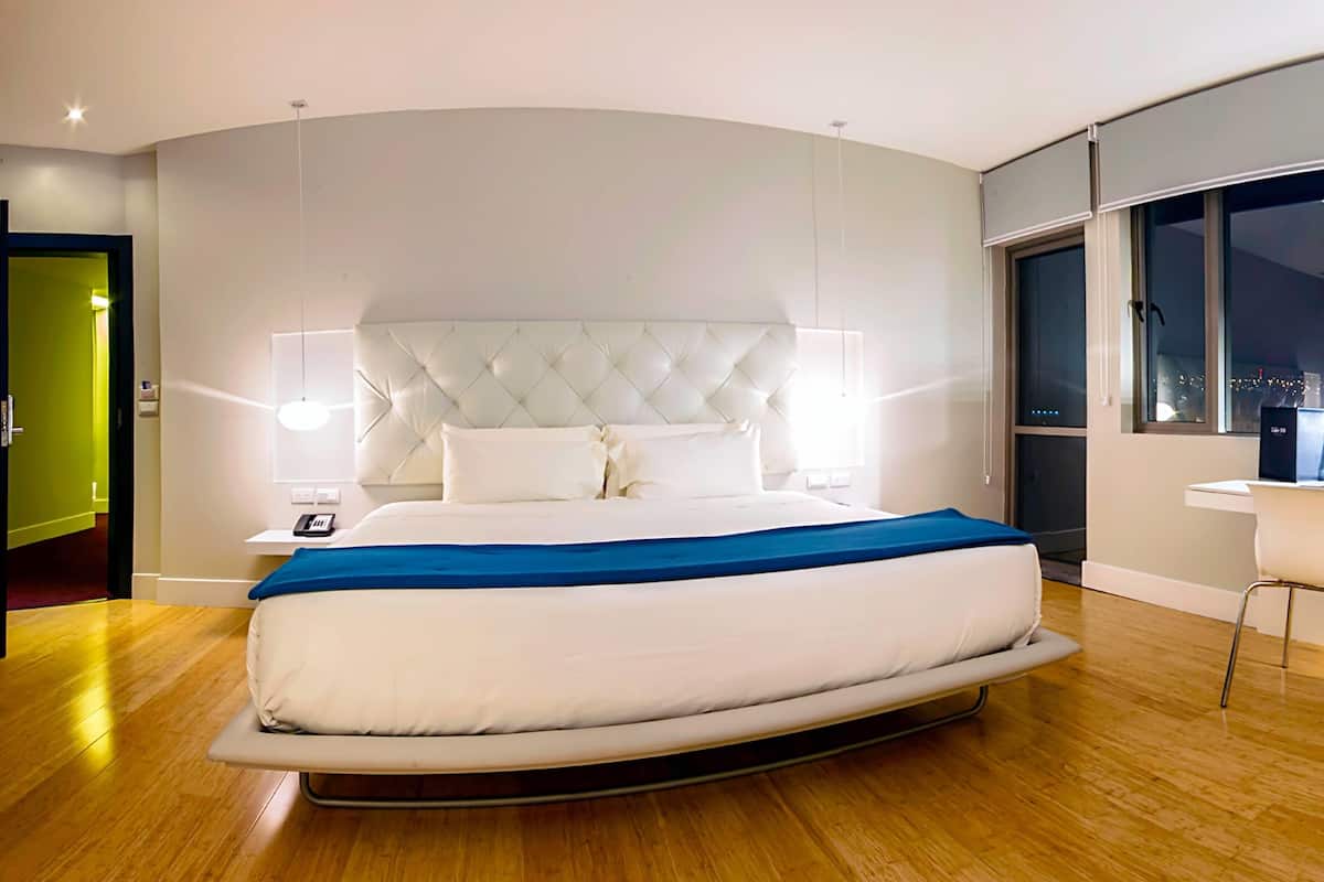 A modern bedroom with a curved bed, a tufted headboard, bedside lamps, and a view of the city at night.