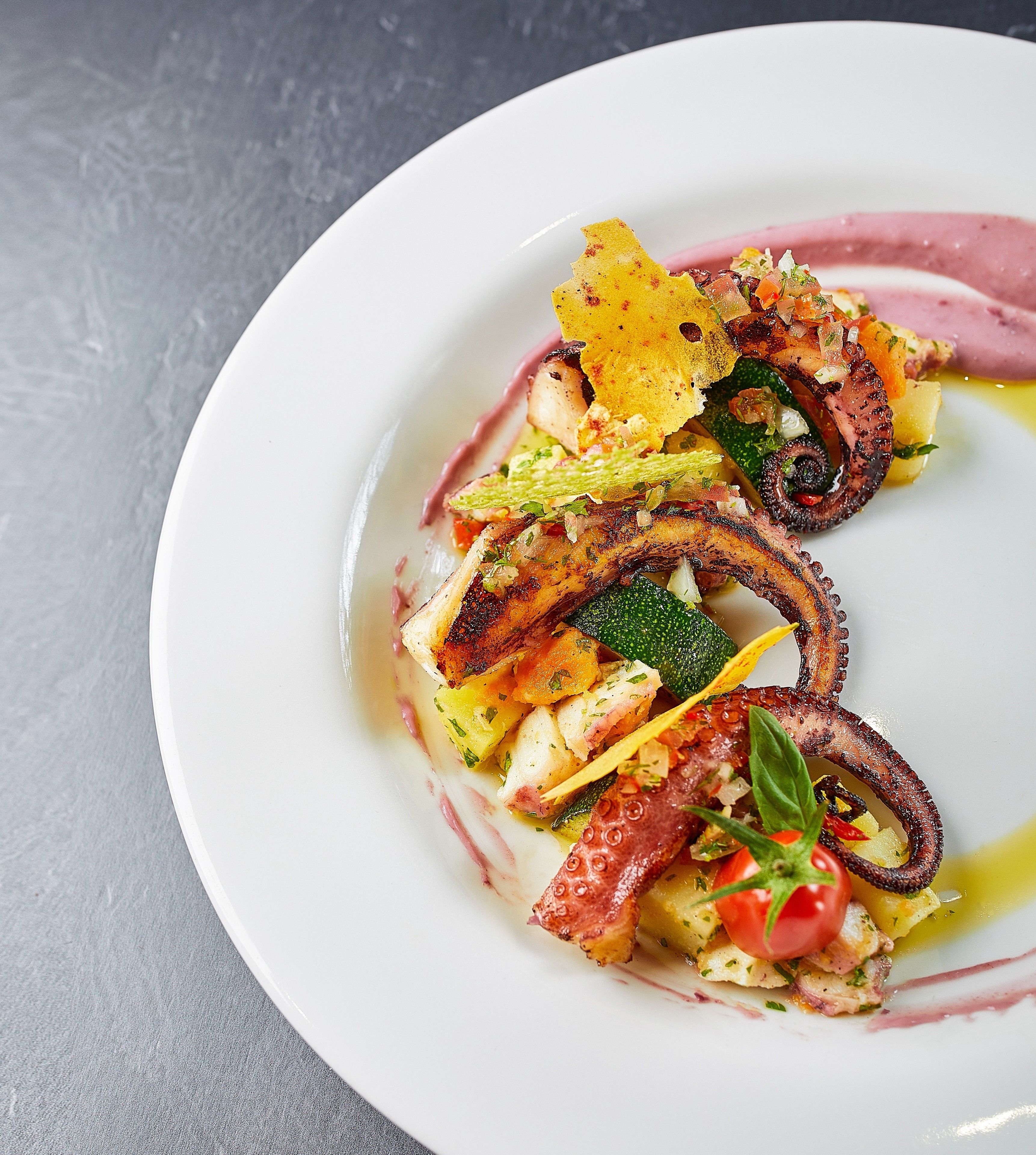 Un plato blanco con diversos alimentos, entre ellos pulpo a la parrilla, verduras y un tomate pequeño.