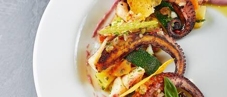 Un plato blanco con diversos alimentos, entre ellos pulpo a la parrilla, verduras y un tomate pequeño.