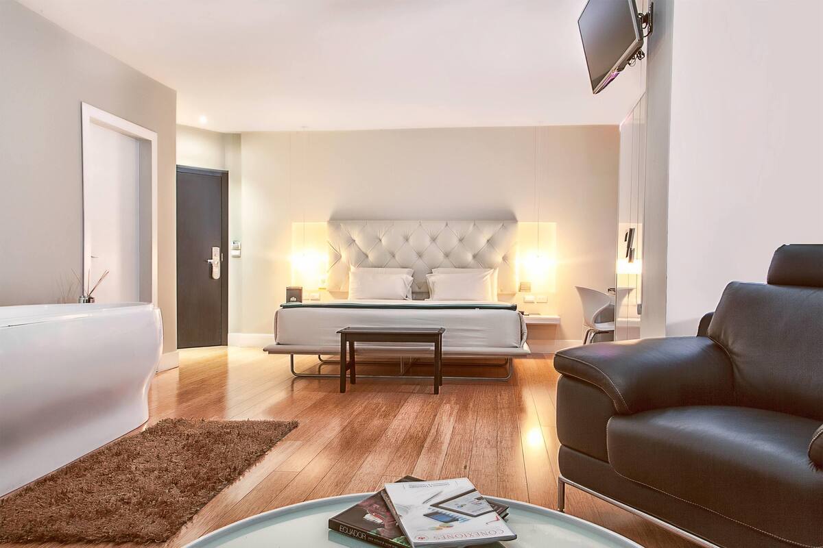 A modern bedroom with a large bed, bedside lamps, a small table, a leather sofa, and a TV mounted on the wall.
