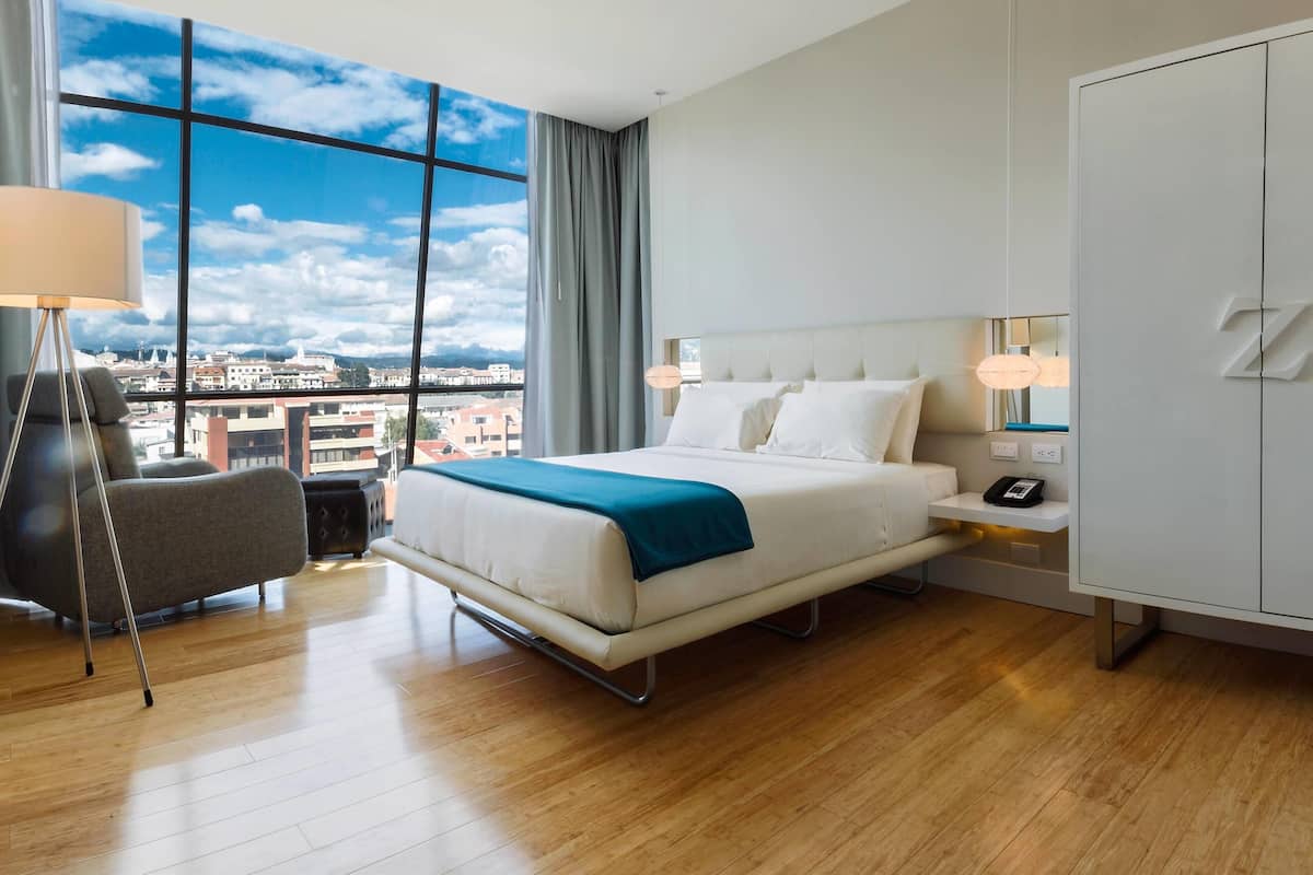A hotel room with a large window, a bed, a bedside table with a phone, a chair, and a wardrobe.