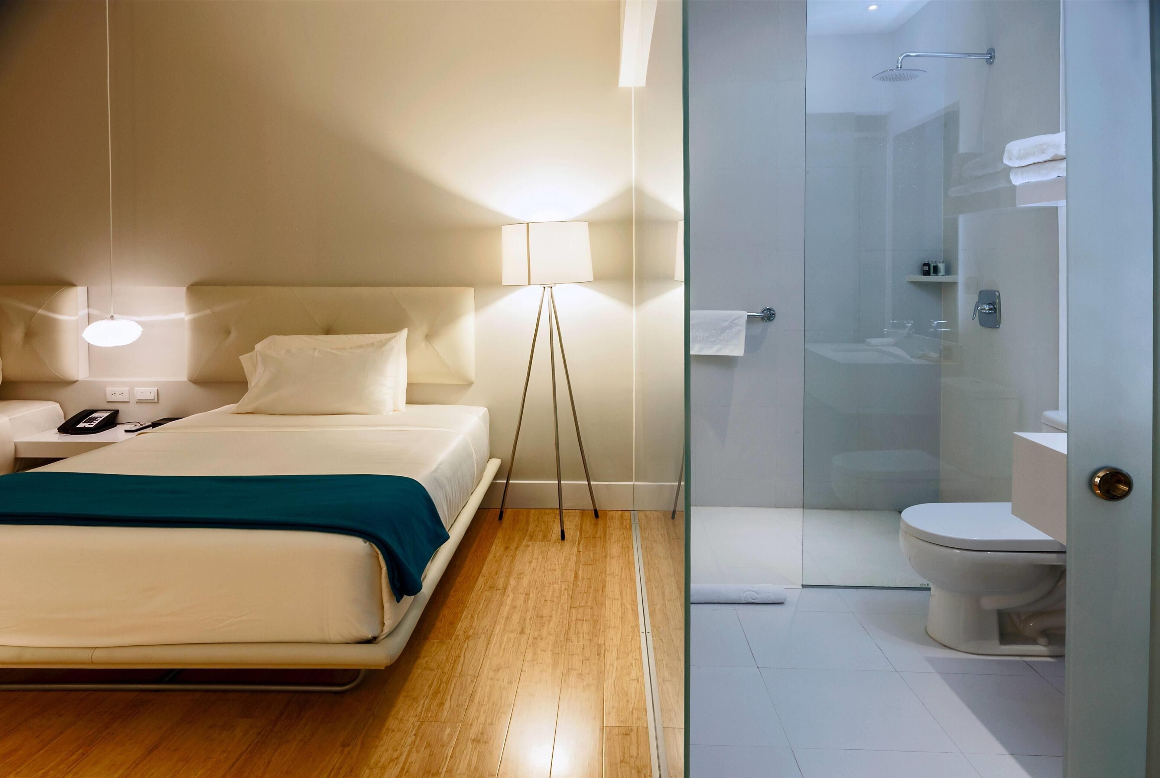 A hotel room with a bed, bedside table, lamp, and a glass-enclosed shower area.