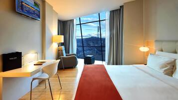 A hotel room with a bed, a desk, a chair, a TV, and a large window with a city view.