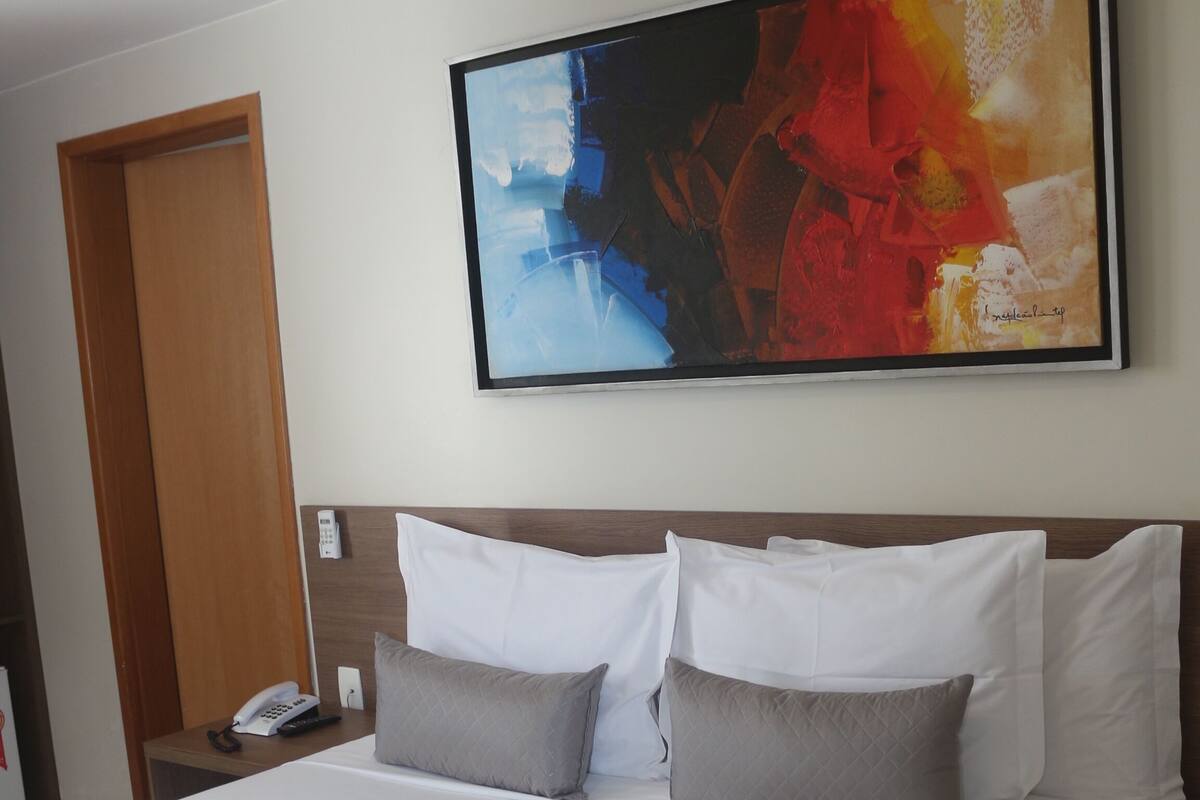 Une chambre d’hôtel avec un grand lit, une table de chevet avec un téléphone et une peinture abstraite aux couleurs vives accrochée au mur.