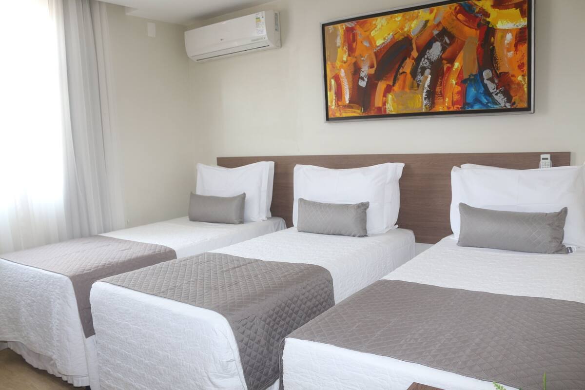 Une chambre d’hôtel avec trois lits, chacun recouvert d’une couverture blanche et grise. Un grand tableau abstrait est accroché au mur, au-dessus des lits.