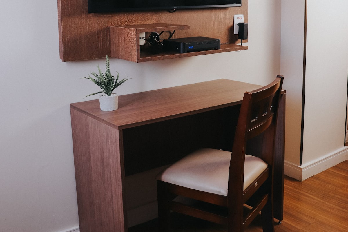 Une pièce de taille modeste, comprenant un bureau en bois, une chaise, un téléviseur à écran plat fixé au mur et une petite plante.