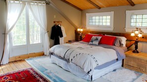 Un dormitorio con una cama grande, dos ventanas, vigas de madera, una mesita de noche con lámpara y un tapete con estampado rojo y blanco.
