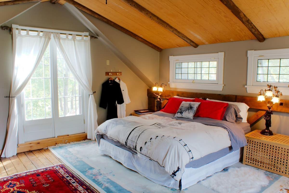 A bedroom with a large bed, two windows, wooden beams, a nightstand with a lamp, and a red and white patterned rug.
