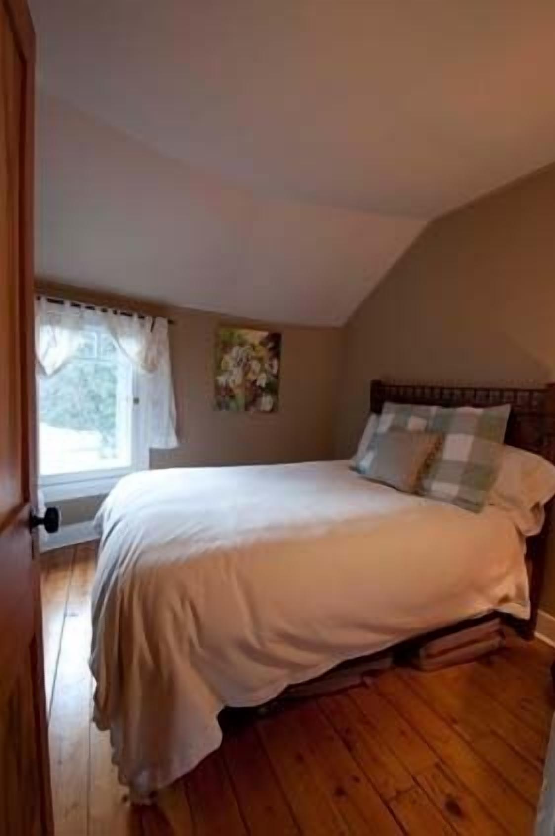 A bedroom with a large bed, wooden flooring, a window with curtains, and a painting on the wall.