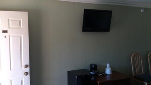 Quarto com porta branca, televisão preta presa na parede, pequena mesa com cafeteira e duas cadeiras de madeira.