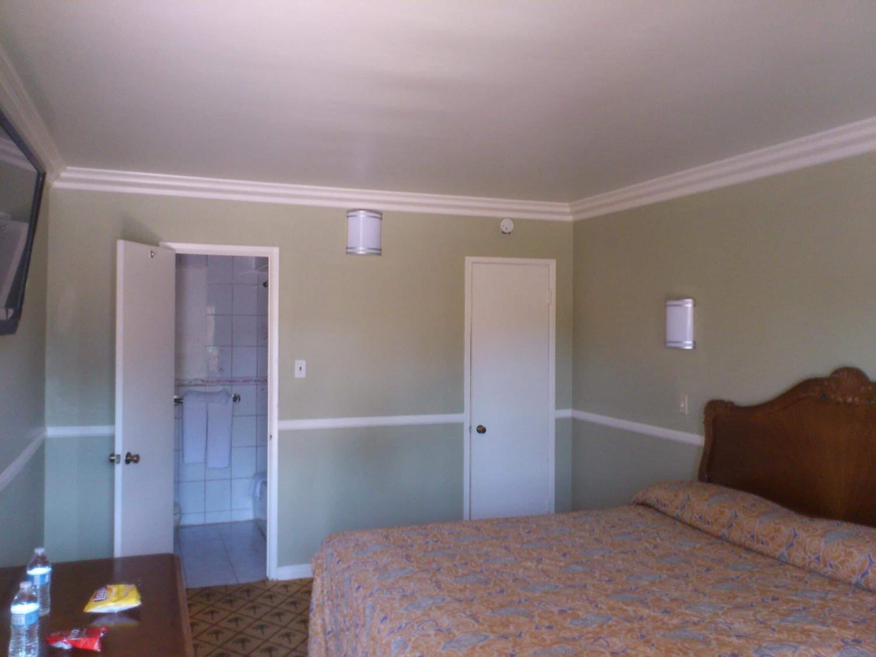 A hotel room with a bed, a table with a bottle and snack, a door leading to a bathroom, and a wall-mounted light fixture.